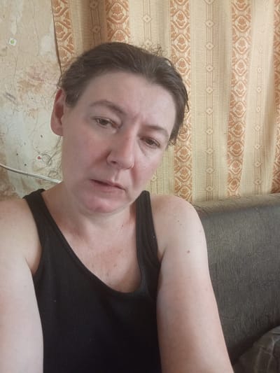 A woman with dark hair pulled back is shown from the chest up, looking downwards with a worried or sad expression. She is wearing a black tank top. She is seated on a dark-colored couch in front of a wall covered with patterned wallpaper featuring vertical stripes and floral motifs in shades of yellow and orange. The environment appears to be indoors, possibly a residential setting. There are no discernible elements indicating the specific location within Berezivka, Ukraine.