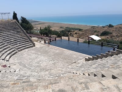 This media file depicts an ancient amphitheater overlooking the Mediterranean Sea in Episkopi, Cyprus. The amphitheater is constructed of stone, with tiered seating carved into the hillside. A vast, dark blue reflecting pool is situated in front of the stage area, reflecting the clear blue sky.

A lone woman in a white dress and black handbag walks across the stone-paved area in front of the pool. She appears to be a tourist, exploring the historical site. The scene is bathed in bright sunlight, suggesting it is midday on a clear, warm day. The landscape surrounding the amphitheater is dry and arid, typical of the Mediterranean climate, with patches of green vegetation and fields stretching towards the coastline. The sea is calm and a vibrant turquoise.

In the background, a white tent structure is visible, possibly providing shade or shelter for visitors. Rows of spotlights are positioned on a platform to the left of the amphitheater's seating. The overall impression is one of historical grandeur combined with a tranquil, scenic natural setting. There is no visible text in the image.
