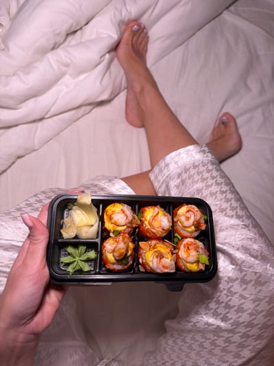 A person in houndstooth pajamas sits in a bed with white sheets, holding a black plastic sushi container. The container has six sushi rolls topped with shrimp and fish eggs, alongside a compartment with ginger and wasabi. The person's bare feet are visible in the periphery. There is no indication of the location being Svkh Voronezhskiy, Russia.