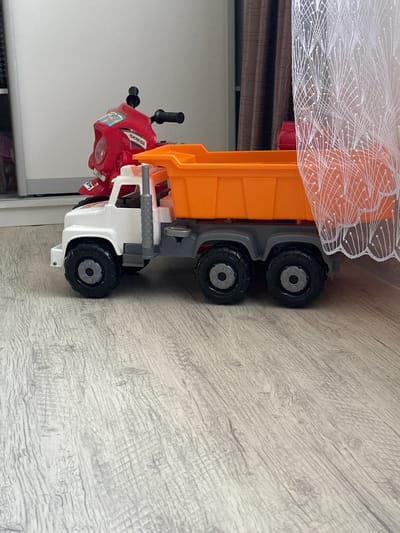 A toy dump truck with an orange bed and a white cab sits on a light-colored, wood-grain floor. Behind the dump truck, a red toy motorcycle is visible. A sheer white curtain with a decorative pattern hangs to the right of the dump truck, obscuring part of the view. The scene appears to be indoors, likely a residential setting, given the presence of children's toys and a curtain. There is no indication of the specific location within the city of Мариинский, Russia.