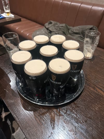 A silver tray laden with full pints of Guinness sits on a dark wooden table. The creamy white heads of the stout are prominently displayed. An empty pint glass rests near the tray, and another empties is closer to the couch. A brown couch is visible in the background, as well as a folded light jacket. The scene suggests a relaxed social gathering.