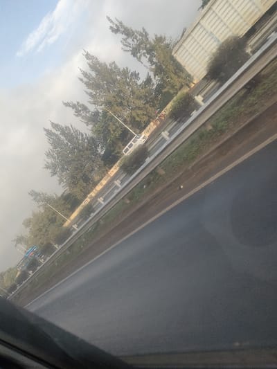 The image depicts a roadside scene in Theta Ward, Kenya, captured from inside a vehicle at an oblique angle. A dark gray asphalt road with a lighter shoulder line occupies the lower half of the frame. A silver metal guardrail separates the road from a green grassy median. Behind the guardrail, a tan concrete barrier displays partially legible text including "PREVENTS INFECT..." and "P from d." A white commercial van is parked or stopped on the far side of the median, oriented away from the viewer. Further left, a blue and white bus stop shelter structure is visible. Numerous tall, green coniferous trees and smaller bushes line the median and roadside, interspersed with streetlights/utility poles. In the upper right background, a large, light-colored industrial-style building or wall is visible. The sky in the upper left corner is cloudy and hazy with some blue patches. No individuals are clearly identifiable in the scene.