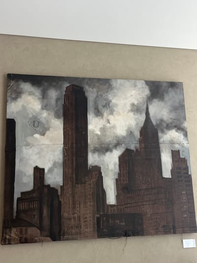 A large, rectangular painting depicting a dark, stylized cityscape is centrally displayed on a light beige textured wall. The artwork, characterized by a muted palette of dark browns, greys, and whites, portrays a dense cluster of tall buildings under a heavily clouded, ominous sky. Prominent features include a slender, dark brown skyscraper in the central-left and a building with a pointed spire-like roof to its right, reminiscent of early 20th-century metropolitan architecture. Visible horizontal and vertical seams across the canvas suggest the artwork may be composed of multiple panels or sections. Staples or fasteners are visible along the upper-left edge of the canvas. A dark, thin wire extends from the lower-right edge of the painting. Below the artwork, a small white label with black text is affixed to the wall. The wall meets a clean white ceiling or trim line at the top of the frame. This scene is located in the city of Galatina, Italy.