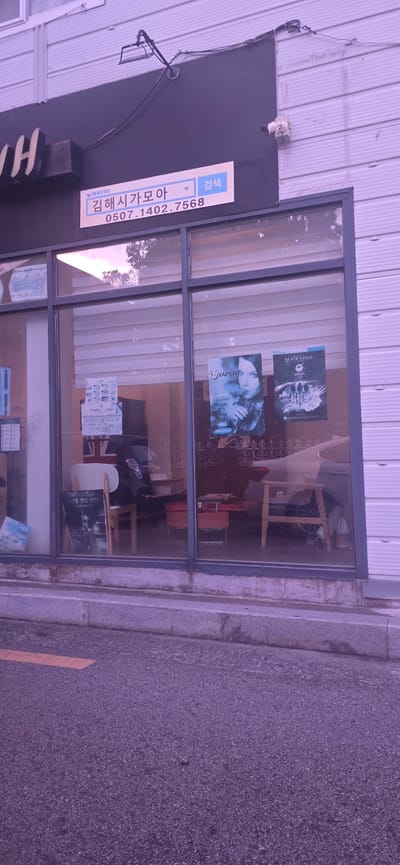 The image shows the exterior of a shop with a large display window. A sign above the window reads "NAVER 김해시가모아 0507.1402.7568" and "검색" (search) in Korean. The display window reveals the interior of the shop, which appears to be a vape shop. Inside, there are posters advertising vape products, including one with the text "VAPER" and another with "BLACK LABEL VERA." There are also chairs, tables, and what appears to be a vehicle visible through the window. The shop is located on a street in Gimhae-si, South Korea, as indicated by the sign. The pavement in front of the shop has a yellow painted line. The building exterior is white and ribbed.