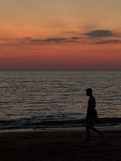 A lone male figure is observed walking from right to left along a dark sand beach in Phra Ae, Thailand. The scene is illuminated by the sunset, casting the individual into a dark silhouette against the brighter background. The man appears shirtless and wears shorts, with a bag or satchel slung over his left shoulder as he walks parallel to the ocean. The body of water occupies the midground, exhibiting a rippled, dark surface with small waves breaking at the shoreline. The sky above presents a vivid gradient of colors, transitioning from light orange and yellow at the top to deep red and purple closer to the horizon, with several elongated dark grey cloud formations interspersed across it. No other individuals or prominent objects are visible in the frame.