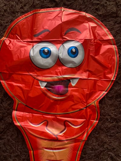 A red, deflated balloon designed to look like a cartoon monster is lying on a dark brown, shaggy carpet. The balloon features a large, round head with two oversized, blue and white cartoon eyes and a wide mouth showing fangs and a pink tongue. Dark, curved eyebrows are drawn above the eyes, and the mouth is outlined in black. The balloon has a distinct shape, tapering down from the circular head to a narrower lower section. The surface of the balloon is wrinkled, indicating it is not inflated. The background is entirely composed of the textured carpet. There are no people or any other objects visible in the image. The scene appears to be indoors, and the lighting suggests it was taken in natural light. There is no information available that can place this image in the city of Mamontovo, Russia.