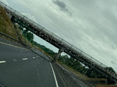 The image, captured from a moving vehicle on a highway, shows a view through a rain-streaked windshield. A concrete overpass, with a metal railing along its edges, spans across the road. The sky is overcast with scattered clouds. The road itself is asphalt, with yellow and white lane markings visible. The surrounding environment consists of green vegetation and trees. The image appears to be taken on the N25 road near Crookstown, County Cork, Ireland.