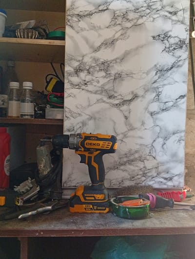 This image captures a cluttered shelf in what appears to be a workshop or storage area. The background features a large sheet with a marbled pattern, predominantly white with black veining. On the shelf, various objects are visible, including a yellow and black cordless drill with the brand "DEKO" clearly visible on its side. Below the drill, an object with "UNV 20V" text is present.
To the left of the drill, there are several bottles with white caps. Further up on a higher shelf, a collection of items including what looks like a woven basket and a green cylindrical object can be seen. A red bottle is also partially visible on the far left. Scattered on the shelf surface are additional tools and items, such as a screwdriver with a red handle and pink grip, a green ashtray-like object containing what seems to be orange material, and other unidentifiable metal objects. The overall setting is a utilitarian space, likely used for storage and DIY projects, as suggested by the presence of the drill and other tools. There are no people or signs of a specific time of day or weather conditions. The location is identified as Suklieia, Moldova.