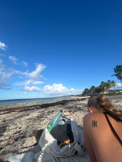 The image displays a sandy beach scene in Le Moule, France, under a clear blue sky with scattered white cumulus clouds. In the foreground, an individual is positioned with their back to the camera, facing the ocean. This individual has blonde hair tied back and wears a dark-colored swimsuit strap on their right shoulder. A dark-colored tattoo, resembling an Adinkra symbol, is present on their left shoulder blade. To the left of the individual, an open light-colored canvas bag rests on the sand, containing folded clothing (dark grey and a multicolored item) and a light-green plastic bottle with a visible "Opes" label. A dark brown sandal is partially visible next to the bag. The beach surface is composed of light-colored sand with patches of dark reddish-brown seaweed at the water's edge. The ocean occupies the mid-ground, featuring calm blue water and a visible reef or rock formation near the shore. Several indistinct figures are visible wading or standing in the water and along the distant shoreline. A line of numerous palm trees extends across the background of the beach, and faint structures are discernible on a elevated terrain beyond the trees.