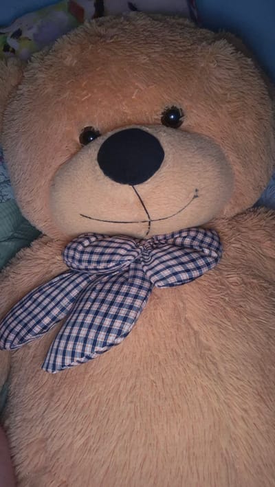 The image shows a close-up, overhead view of a large, plush teddy bear lying on a surface that appears to be a bed or sofa, likely indoors. The teddy bear is a warm, peachy-orange color with a fluffy texture. It is adorned with a bow made of blue and white checkered fabric, tied between its ears. The bear's face is visible, featuring a large, black circular nose, two dark, shiny eyes, and stitched details for its mouth. The lighting suggests an indoor setting, potentially in the evening or during a dimly lit day, as there are no strong shadows. The background is out of focus, but hints of colorful fabric or bedding can be seen. There is no visible text in the image. The overall impression is one of comfort and a relaxed, domestic scene.