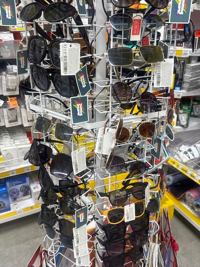 The image shows a display rack filled with sunglasses in a retail setting, likely a store. The sunglasses are of various styles, colors, and lens tints, with many featuring price tags and branding information. Some of the sunglasses are hanging from the rack, while others are stacked. The background reveals shelves stocked with other merchandise, including what appear to be boxes of electronic devices or accessories. A prominent yellow display shelf is visible in the upper right corner. The lighting suggests an indoor environment, possibly a well-lit store. Several labels are visible, including ones that say "Sunglasses" and "Children sunglasses," along with some with what appears to be product information and certifications like "CE." Some of the tags also indicate UV protection up to 400. The overall scene depicts a retail display of eyewear.