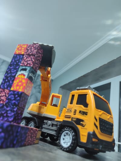A toy excavator truck, predominantly yellow with black and grey accents, is positioned on a dark, textured surface. The truck has a prominent orange arm and a black bucket. To the left of the truck, a tall, colorful structure made of blocks is stacked, exhibiting a pixelated pattern in shades of purple, blue, yellow, and red. The background shows a ceiling with white molding and a hint of a doorway or window. The lighting appears to be artificial, casting some reflections on the ceiling. There are no people visible. The image is a close-up shot, focusing on the toys and their arrangement. Text visible on the truck includes "KING" and some other unreadable symbols and numbers. The scene depicts a playful arrangement of toys, possibly for a child's play area.
