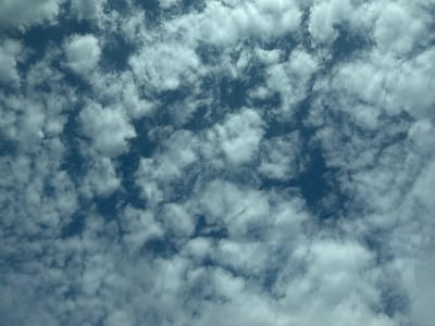 This is a photograph of the sky with scattered cumulus clouds against a blue background. The image is taken from a low angle, looking upwards. There are no people, subjects, or objects visible in the scene, except for the clouds. The environment is an outdoor setting, likely a clear day with some cloud cover. The photograph is devoid of any specific landmarks or context that would allow for precise location identification within the city of Ruma Municipality, Serbia.