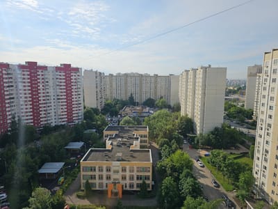 This is an aerial view of a residential area in Khimki, Russia, likely taken from a tall building. The scene is dominated by numerous high-rise apartment buildings, with a mix of architectural styles. On the left, a large, striking red and white apartment building stands out. To its right and in the center of the image are several light-colored, long, rectangular apartment blocks, characteristic of Soviet-era architecture. These buildings are arranged in a way that creates courtyards and open spaces.
Interspersed between the buildings are dense clusters of mature green trees, indicating a well-established urban green space. Several smaller, lower-rise buildings, some appearing to be schools or kindergartens with playgrounds, are visible nestled within the trees. Roads wind through the complex, with cars parked along the sides and some driving on them, suggesting normal daily activity. The sky is blue with scattered white clouds, and the sunlight casts shadows, indicating it is daytime. In the distance, more buildings and urban sprawl are visible, characteristic of a large city. Power lines crisscross the sky above the buildings.