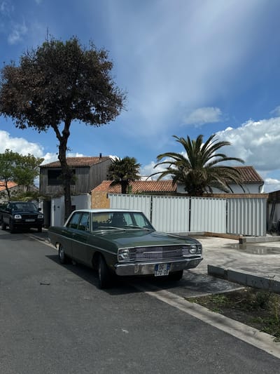 The image shows a residential street scene in La Couarde-sur-Mer, France. A vintage green car is parked on the right side of the road, facing away from the viewer. The car's license plate, "DG-670-DB," is visible. To the left of the car is a white fence, behind which are buildings with tiled roofs and a palm tree. Further down the street is a dark-colored SUV. A large tree with sparse foliage is located on the left side of the street, under a blue sky with scattered clouds. The road surface is asphalt.