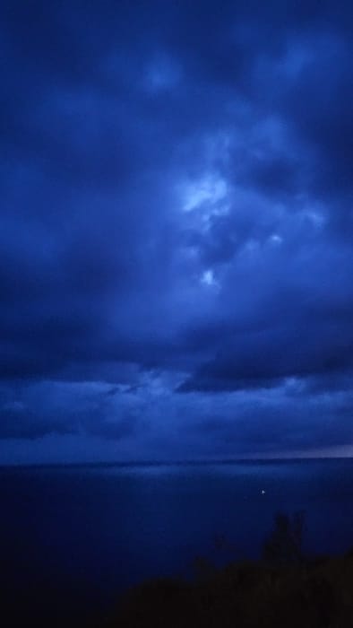 The image depicts a coastal vista at night or late dusk from an elevated position in Santa Ponça, Spain. The upper two-thirds of the frame consists of a dark blue, overcast sky filled with dense clouds. Several localized areas within the cloud mass show brighter, lighter blue illumination. A defined horizon line separates the sky from the body of water below. The lower third of the image shows a calm, dark blue expanse of water. A single, distinct white light is present on the water's surface, positioned slightly right of the frame's vertical center. The bottom edge of the image reveals dark, indistinct silhouettes consistent with terrestrial foliage or terrain. No people, discernible actions, or interactions are visible.