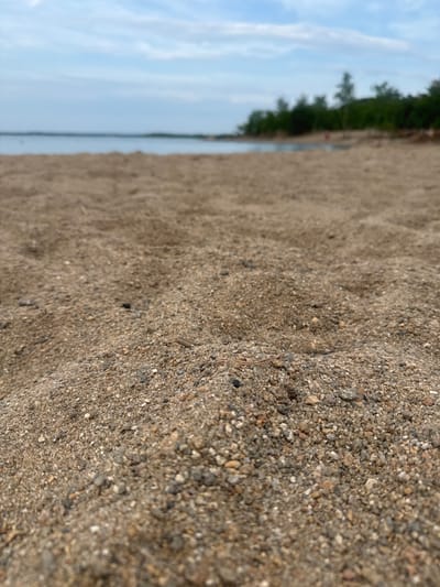 The image depicts a close-up view of sandy ground, with a body of water and trees visible in the background. The sand is a mixture of fine grains and small pebbles. The water appears calm, and the trees are dark green, suggesting they are evergreen or have dense foliage. The sky is a pale blue with some wispy clouds, indicating it is daytime, likely overcast or hazy. The perspective is low to the ground, focusing on the texture of the sand. There are no people or distinct objects clearly visible in the foreground or midground. The overall scene evokes a natural, outdoor setting, possibly a beach or lakeshore. No text is visible in the image. The location context provided is Володарский ауылдық округі, Kazakhstan.