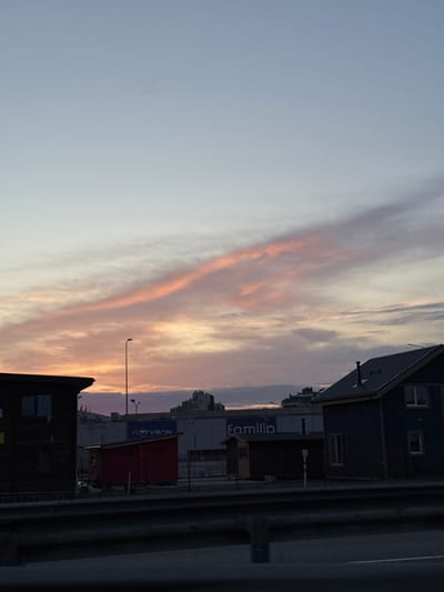The image shows a street scene in Lappeenranta, Finland, at dusk. In the foreground, there is a dark highway barrier. Behind the barrier, there are several buildings, including a dark-colored house on the right with a dark roof and a window visible. To the left of this house is a smaller dark-colored shed. Further to the left, there is a red shed. In the background, there is a large building with a sign that reads "familia". Above the buildings, the sky is a gradient of pale blue and pink, with wispy clouds illuminated by the setting sun. There are also streetlights visible in the scene. The overall impression is a quiet, urban landscape at the end of the day.