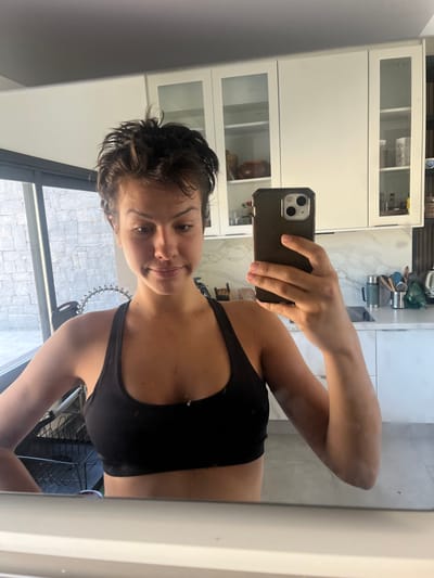 A female individual is captured in a mirror selfie within a modern residential kitchen in La Barra, Uruguay. The subject has short, dark, tousled hair and is wearing a black sports bra. She holds a dark-cased smartphone, consistent with an iPhone model featuring a multi-lens camera system, in her right hand to photograph her reflection. Her gaze is directed slightly downwards and to her right, displaying a subtle, neutral expression.
The background reveals a contemporary kitchen environment. On the right, white upper cabinets, some with frosted glass inserts, are mounted above a light-colored stone or marble countertop. White lower cabinets are visible beneath the counter. Kitchen items on the countertop include a metal thermos, a black utensil holder containing various utensils, and a white electric kettle. A dark, vertically slatted panel is present on the far right. To the left of the subject, a large window or glass door overlooks an exterior area with a textured grey stone wall. A chrome flexible faucet hose and a black dish drying rack are partially visible near the bottom left of the window area. The scene is brightly illuminated, indicating the presence of natural light from the window and ambient interior lighting.