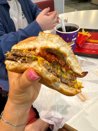 A person with pink nail polish holds a halved double cheeseburger. Visible burger ingredients include two beef patties, melted cheese, bacon, pickles, and ketchup. A McDonald's french fry container and a McDonald's drink cup with a straw are in the background. The location is Spreitenbach, Switzerland, identifiable by the McDonald's packaging. In the bottom left corner, a child's hand rests on a red surface. A person's leg and a silver chain bracelet on their wrist are visible in the lower right. Crumpled fast-food wrapping paper is in the background near the burger.