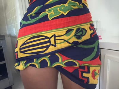 A person is wrapped in a colorful sarong featuring red, yellow, green, and blue patterns. The fabric appears to have designs of flowers, vases and geometrical shapes. The person's upper thighs and stomach area are visible, but other parts are obscured by the fabric. The setting is indoors, close to white cabinets and a light-colored tiled wall. A small, clear container with pink objects inside is affixed to the cabinet's side.
