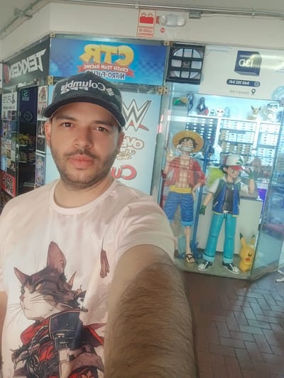 A man takes a selfie inside a brightly lit retail establishment in Lince, Peru, specializing in video games and pop culture merchandise. He is positioned in the foreground, looking directly at the camera with a neutral expression. He wears a dark baseball cap with "Columbia" written on it (appearing mirrored in the image) and a light-colored t-shirt featuring a detailed graphic of a cat, possibly in a samurai or warrior-like outfit.
Behind the man, a large display case or shop window is visible, showcasing prominent anime figures. To the left, a tall figure of Monkey D. Luffy from One Piece stands, while to the right, Ash Ketchum from Pokémon is displayed, with a smaller Pikachu figure at his feet. Above these figures, various smaller collectibles are arranged on shelves.
The background reveals several signs and posters related to gaming and entertainment. Above the display case, a large banner is visible, with mirrored text clearly showing "CTR" and "CRASH TEAM RACING NITRO-FUELED". Another sign features the "WWE" (World Wrestling Entertainment) logo. To the far left, a shelf or stand displays various game titles, including "TEKKEN" and references to "PS5". An emergency light fixture is mounted on the ceiling, with the mirrored Spanish text "LUCES DE EMERGENCIA" (Emergency Lights) visible. Another mirrored sign on the right displays "13D" and what appears to be partial address information like "Stand 00-1". The floor is tiled with reddish-brown bricks.