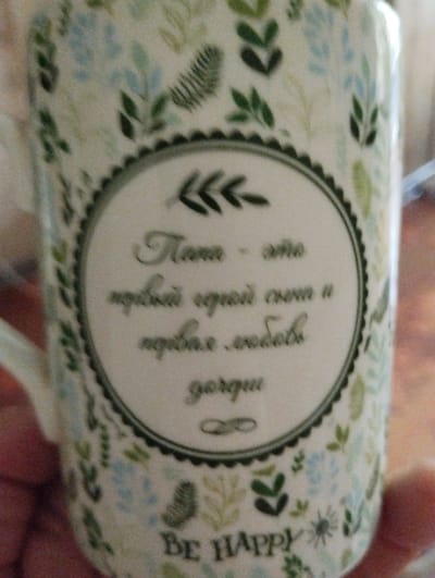 The image shows a close-up of a ceramic mug with a floral pattern and text. The mug is decorated with light green and blue leaves and small blue flowers on a cream-colored background. A dark green, circular border with a serrated edge encloses the text. The text appears to be in Russian. The first line reads "Папа -" which translates to "Dad -". The second line is "это лучший серый сон и" which translates to "is the best grey dream and". The third line is "первая любовь" which translates to "first love". The fourth line is "дорогие" which translates to "dear" or "darling". Below the circular border, the text "BE HAPPY" is visible. A hand is holding the mug from the right side of the image. The background is blurred and out of focus, suggesting the mug is the primary subject. There is no information in the image to suggest its location in the city of Sheptytskyi, Ukraine.
