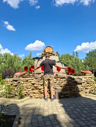 A man in a black t-shirt and khaki shorts stands with his arms outstretched in front of a stone structure decorated with large wine glasses filled with red liquid. The structure, which resembles a wine barrel, is adorned with clay pots and a wooden barrel on top. Behind the structure, lush green trees and a bright blue sky with white clouds form the background. The scene appears to be outdoors, possibly in a winery or tourist attraction. The man is positioned in the foreground, facing away from the viewer, with his shadow cast on the paved ground. The location is identified as Milestii Mici, Moldova.