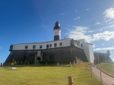 The image displays the Forte de Santo Antônio da Barra and the Farol da Barra (Barra Lighthouse) in Salvador, Bahia, Brazil, under a bright blue sky with scattered white cumulus clouds. Intense sunlight originating from the right side of the frame illuminates the scene, creating minor lens flare and distinct shadows.
The central subject is a historic fortress, characterized by a substantial stone base with white-painted upper battlements. The fort structure includes multiple small rectangular windows and defensive bastions at its corners. A prominent arched entrance, centrally located on the main facade, is topped by an ornate, multicolored heraldic crest. Rising directly from the fort's structure is the Farol da Barra lighthouse, a tall cylindrical tower with alternating black and white horizontal stripes, culminating in a dark-colored lantern room.
The foreground features an expansive, well-maintained green lawn that gently slopes upward towards the fort. On the left side of this lawn, a group of approximately six individuals, some wearing yellow high-visibility clothing, are gathered near a piece of yellow machinery, indicating activity such as construction or maintenance. Two small, white rectangular concrete blocks are also present on the grass in this area.
On the right side of the image, a paved pathway, constructed from reddish-brown pavers, curves and ascends towards the fort. This path is bordered by a series of temporary white posts interconnected by orange tape or string. Approximately four to five individuals are observed walking uphill on this path towards the fort. Additional people are visible at the fort's main entrance, either entering or exiting the structure.