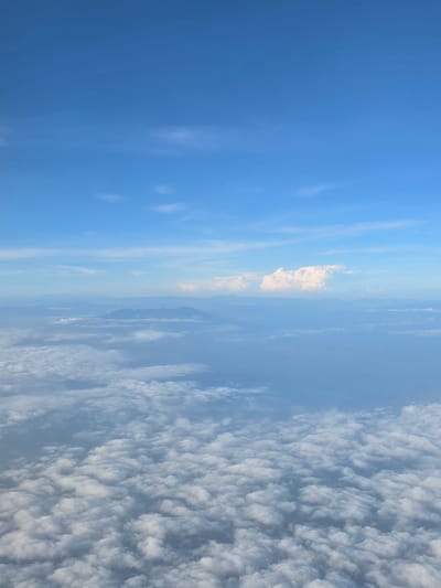 The image captures an aerial view of a vast expanse of clouds, with a clear blue sky above. In the distance, a range of mountains is faintly visible through a haze. The scene suggests a flight over Indonesia, given the reference to Tanohanoe. There are no people or man-made structures visible in the image, only natural elements of the sky and landscape. The overall impression is one of serene natural beauty.