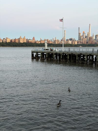 The image shows a view of the New York City skyline across the Hudson River from Edgewater, New Jersey. In the foreground, two Canada geese swim in the choppy water. A pier extends into the river from the left side of the frame, with people sitting on chairs at the end of it. A statue of a person is located on the pier, and an American flag flies from a flagpole. The background features a dense line of trees and a collection of buildings, including residential apartment buildings and skyscrapers, with the distinct architectural styles of Manhattan visible. The sky is a soft gradient of pale blue and white, suggesting it is either early morning or late evening.
