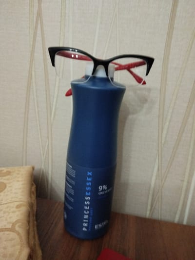 The image shows a pair of black-rimmed glasses with red accents resting on top of a blue bottle with white text. The bottle appears to be a cosmetic product, possibly hair dye, with text visible that reads "PRINCESSESSEX" and "9% OXYCHED". The bottle is standing upright on a dark wooden surface, possibly a table or shelf. In the background, a wall with vertical stripes of a light beige color is visible. To the left of the bottle, a portion of a cushioned object, likely a pillow or a piece of furniture, is seen. The overall setting suggests an indoor environment. There are no people present in the image. The image was taken in Kamensk-Shakhtinsky, Russia, and depicts a still life arrangement of personal items.