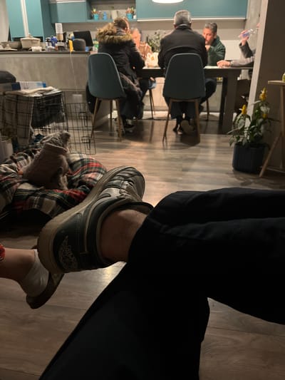 The scene is in an interior space. In the background, there are several people seated at tables, seemingly engaged in conversation or a meal. The foreground features a person with their legs crossed, wearing black jeans and a flannel blanket. One foot is in focus, wearing a gray sneaker with the Air Jordan logo visible on the back. The sneaker appears worn. The flooring is wood. There is a room visible in the back with blue walls. The location is unspecified within Fraccionamientos del Sur, Mexico.