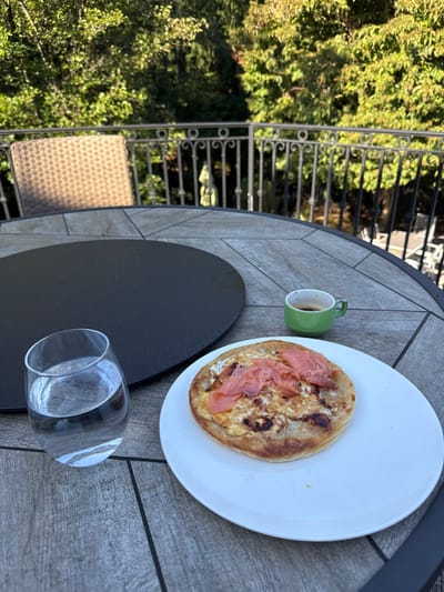 The scene depicts an outdoor patio or balcony in Cresskill, United States, viewed from a slightly elevated perspective. A round outdoor table with a gray wood-look tiled surface and a dark circular insert in its center occupies the foreground and midground. On the table, a white oval plate holds a meal consisting of a round, flatbread-like base topped with cooked egg, a white creamy spread, and multiple slices of smoked salmon. To the right of the plate, a small green ceramic cup contains a dark beverage, likely coffee. To the left, a clear, stemless glass is filled with water. A brown wicker-style chair is partially visible in the left background. A dark metal railing with decorative scrollwork spans across the midground, separating the patio from a dense expanse of green trees under natural daylight. No individuals are present in the image.