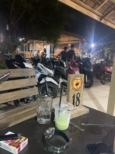 The image shows a table on the street in Gampongaceh, Indonesia, with a green smoothie in a plastic cup, a glass, a watch, a pack of cigarettes, and a lighter. Several motorcycles are parked near the table. People are sitting at tables in the background. It's nighttime.