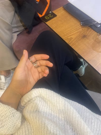 The image depicts an indoor scene in Monastir, Tunisia, from an overhead perspective, focusing on the immediate surroundings of a seated individual. The foreground features a light-skinned right hand resting, adorned with a plain band ring on the ring finger and an ornate, possibly snake-like, ring on the middle finger. The hand is positioned over the lap of the individual, who is wearing a textured, cream-colored knit sweater and dark, possibly black, trousers. To the right, a light-toned wooden table surface is visible, featuring a black table number stand with "29" displayed in orange, positioned upside down from the camera's view. On the table, a white ceramic plate is stacked over a bundle of cutlery (fork and knife) wrapped in a napkin, alongside a clear drinking glass containing a transparent liquid. To the left, a portion of another person's leg is visible, clad in light-colored trousers and a white sneaker with dark accents. The background includes blurred purple-hued upholstered seating and a partial view of an orange and black object, possibly a bag strap, behind the table number.