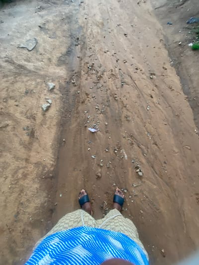 The image depicts a first-person perspective of a person standing on a dirt path in Jere South, Nigeria. The path is wet and appears to have been recently rained on, with visible water channels and debris scattered across the surface, including small rocks and what looks like a piece of plastic. The person is wearing black sandals and a patterned garment, possibly shorts or a skirt, with a blue and white striped fabric draped over their lap. The environment is natural and appears to be an outdoor area, with hints of green vegetation in the upper right corner. The scene suggests a simple, perhaps rural, setting within the specified location.