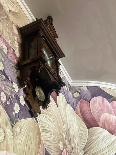 An ornate, dark brown wooden wall clock is mounted on a floral-patterned wall in Dmitriyevskaya, Russia. The clock features a carved pediment at the top, decorative moldings along its structure, and a glass-encased face with light-colored numerals and hands. Below the main clock face, a circular, metallic, embossed disk is visible, likely a pendulum bob. The wall is covered with wallpaper depicting large-scale floral designs in shades of purple, beige, and pink, with smaller white floral elements. A smooth, light-colored ceiling meets the wall via white crown molding, visible in the upper right portion of the frame. The image is captured from a low-angle perspective, looking upwards towards the clock and the ceiling. No individuals or discernible actions are present.