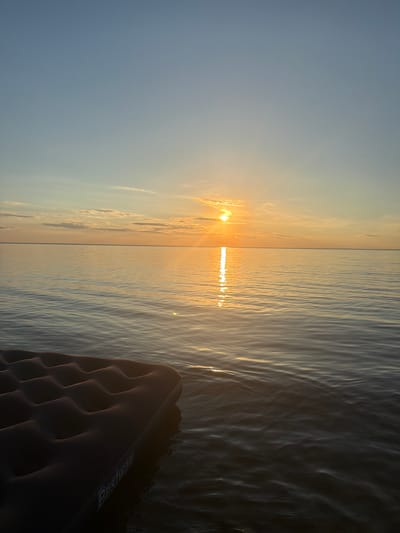 An inflatable mattress, with the "Bestway" logo visible, floats on calm water under a clear sky. The sun is setting on the horizon, casting a warm orange glow and a reflection on the water's surface. The scene is set at the Black Sea coast in Lebedivka, Ukraine.
