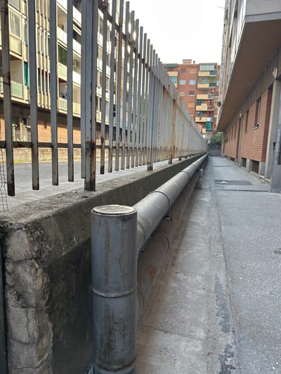 The image depicts an outdoor scene in the city of Trieste, Italy. In the foreground, a concrete wall runs along the left side, showing signs of wear and tear. Resting on this wall are two large, parallel pipes, one above the other, made of weathered metal. The upper pipe is grey and has circular clamps. The lower pipe is darker and shows patches of rust. To the right of the pipes, a paved walkway recedes into the background. Along the left edge of the image, a tall, metallic fence with vertical bars is visible. Beyond the fence, a multi-story building with a light-colored facade and numerous windows can be seen. Further in the background, other buildings with balconies and awnings are present. The sky is overcast. The scene appears to be a utilitarian area, possibly an alleyway or service passage between buildings, characterized by industrial elements and urban infrastructure.