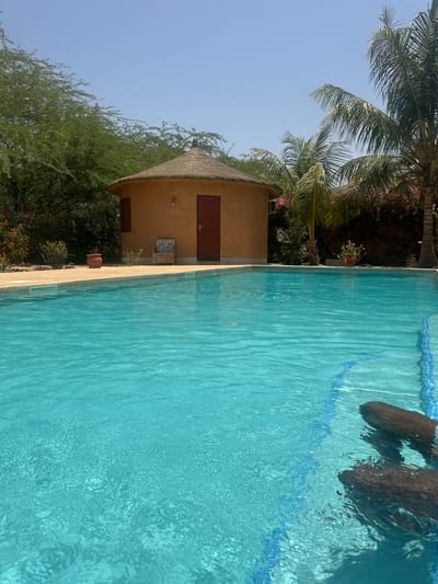 The image shows a swimming pool adjacent to a round, thatched-roof building with a brown door and a small window. The building is surrounded by trees and plants. In the pool, there is a glimpse of what could be feet under the water. The location appears to be Communauté rurale de Malicounda, Senegal.