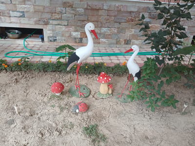 The image depicts an outdoor scene in the city of Yukariyuz, Uzbekistan. In the foreground, the dry, cracked earth is decorated with several garden ornaments. These include two large stork statues, one on the left and one on the right, both white with black wings and red legs and beaks. Between the storks are two red and white mushroom-shaped ornaments, one larger and one smaller, and a ladybug ornament painted on a stone. The middle ground features a row of green foliage with small orange flowers. In the background, a house wall constructed of various colored bricks is visible. A green garden hose is laid out on a patterned tiled walkway in front of the wall. A metal basin and what appears to be gardening tools are visible within an open recess in the wall. The overall environment appears to be a well-maintained garden or yard area.