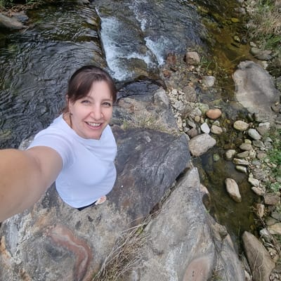 In the city of Mureungdowon-myeon, South Korea, a woman takes a selfie from a high vantage point overlooking a shallow stream flowing over rocks. She is positioned on a large, flat rock formation, with her body angled towards the camera, her right arm extended to capture the shot. She has brown hair pulled back and is wearing a white t-shirt. Her expression is a wide smile, revealing her teeth.
The stream is clear and shallow, with visible rocks on its bed and banks. The surrounding environment consists of more rocks of various sizes and some dry grass. The water appears to be in a natural setting, likely a park or outdoor recreational area. The overall impression is one of a spontaneous moment captured during an outdoor excursion.