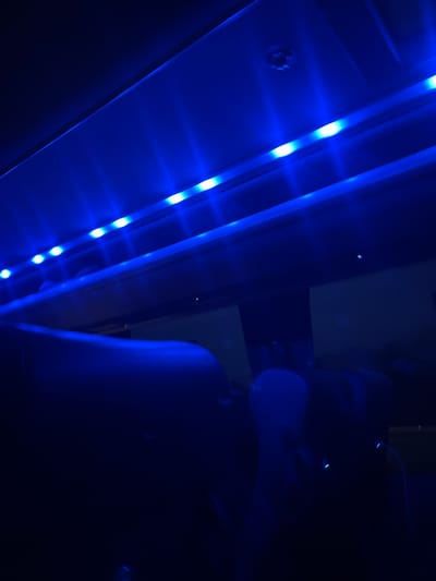 The image captures a dimly lit indoor scene dominated by intense blue light from LED strip lighting. A prominent row of individual blue LED bulbs runs diagonally across the left side of the frame, affixed to a lighter-colored panel. These lights emit bright, focused beams, illuminating the immediate surroundings and casting a pervasive blue glow over the entire photograph.
To the far left, a small, dark metallic, circular fixture, possibly a screw or bolt head, is partially visible. On the right side, a large, indistinct, dark blue shape suggests a piece of furniture, such as an armrest or part of a chair, rendered almost silhouette-like against the dark background due to the low light. The background overall is very dark, bordering on black, with subtle hints of other structural elements or room features.
The setting appears to be an interior space, likely a room or possibly a vehicle cabin, given the nature of the accent lighting. The strong artificial illumination from the LEDs and the deep shadows indicate that the image was taken at night or in a very dark environment. There are no people visible, nor is any specific activity taking place. The blue lighting creates a cool and modern, perhaps atmospheric, ambiance. No discernible text is present in the image. The location context of Woking, United Kingdom, is not visually evident from the scene.
