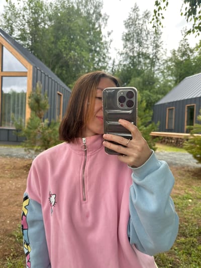 A young woman takes a selfie with her phone in Novaya Vozhoika, Russia. She is wearing a pink and blue sweatshirt with a Hello Kitty graphic. Behind her, two modern-style cabins with dark siding and metal roofs are visible, surrounded by trees and a gravelly area. The sky is overcast.