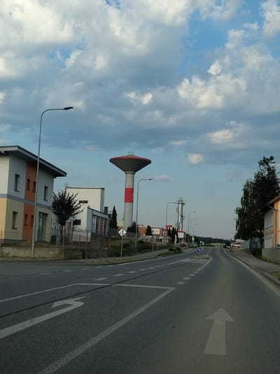 This media file captures a view of a street scene in Horažďovice, Czechia, with a prominent water tower. The camera is positioned at an angle, giving a slightly tilted perspective of the surroundings. The sky is filled with a mix of white and gray clouds, suggesting a partly cloudy day.
The main subjects include a tall, red and white striped water tower with a dark conical top. Beside it, several buildings are visible, some appearing to be industrial or commercial with modern architectural designs. One building has a facade with alternating bands of gray, orange, and white. A metal fence encloses some of these properties.
A road runs through the scene, marked with white directional arrows and lane dividers. The road appears to be an asphalt surface. Streetlights are also visible along the side of the road. In the background, trees and greenery can be seen, indicating an outdoor setting likely during the daytime, possibly late afternoon or early evening due to the lighting. There is no discernible activity of people taking place, and the scene conveys a sense of quiet or stillness. No text is clearly legible in the image.