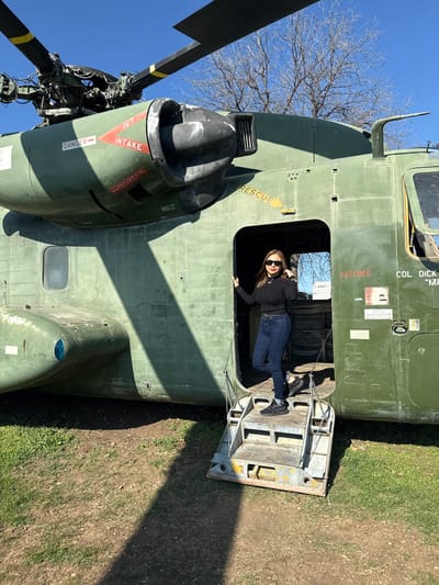 A woman in blue jeans, black shoes, and a black long-sleeved shirt poses in the doorway of a military helicopter. The helicopter is olive green and appears to be a decommissioned aircraft displayed on a patch of grass and dirt. The helicopter door is open, providing a view of the interior. Part of the rotor and a tree are also visible. The sky is a clear, bright blue.