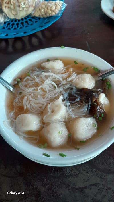 A close-up shot captures a bowl of fish ball soup with noodles and dark mushrooms. The soup is served in a white bowl and garnished with chopped green onions. To the upper left of the bowl, a blue woven basket contains packets of snacks, possibly peanuts and crackers. The background is a dark, wooden surface. The image has a watermark in the bottom left corner that reads "Galaxy A13". The scene appears to be indoors.