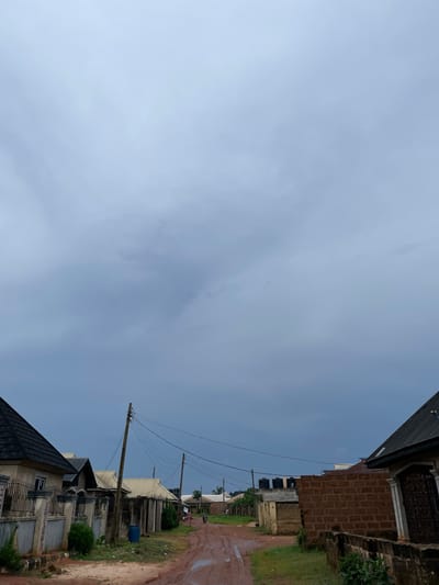 The image depicts a cloudy sky over a street in Edopa, Nigeria. On the left, houses with black and light-colored roofs and metal fences line the dirt road. A utility pole with multiple wires stands in the middle of the road. The road itself is wet, with visible puddles, suggesting recent rainfall. Further down the street, more houses and fences are visible, along with some vegetation and trees. Two people are walking on the road, and a motorcycle is also present. A blue plastic barrel is located on the side of the road. On the right side of the image, a brick wall and a house with a black roof are visible.