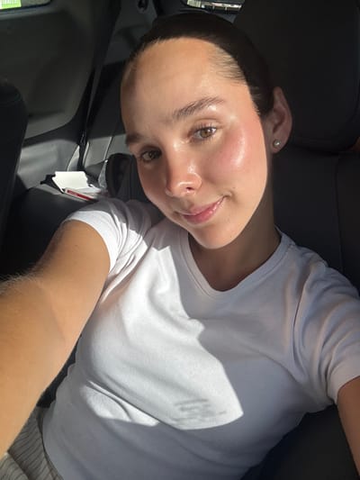 A female individual is depicted in a self-portrait taken inside a vehicle, situated in Ciudad Victoria, Mexico. The subject, a young adult with dark hair pulled back and a light complexion, wears a white short-sleeved t-shirt and a single light-colored stud earring in her left ear. She faces the camera with a slight smile. Direct sunlight illuminates her front, casting a sharp shadow across her upper torso. The background reveals dark-toned vehicle interior elements, including rear passenger seats. A white rectangular container with red markings is visible on the rear seat behind the subject. No other individuals are present within the frame.